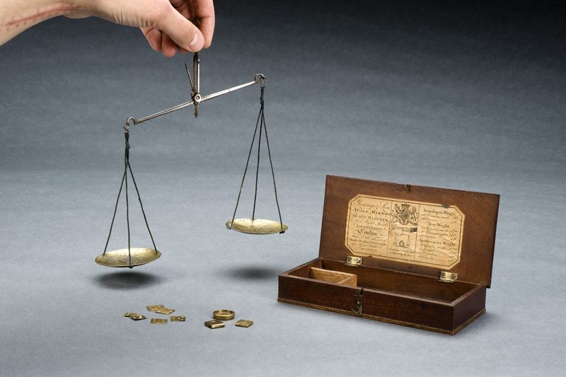A hand holding a small brass balance above its wooden case and weights